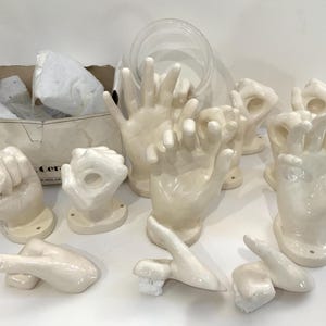 May include: A collection of off-white ceramic hand-shaped decorative objects. The hands are in various gestures, including fists, open palms, and the 'okay' sign. Some are mounted on small circular bases. A cardboard box with packing material is in the background.