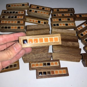 May include: A selection of small, rectangular wooden boxes with mosaic tile detailing. Each box has a series of orange and black square tiles set within a wooden frame. The boxes are arranged on a white surface, with one box being held by a hand.