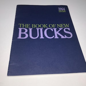 May include: A vintage book titled "The Book of New Buicks" with a dark blue cover. The title is in green and purple lettering. The top right corner reads "1964 Edition". The book is lying on a white surface.