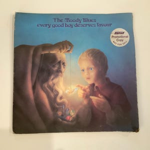 May include: Album cover for The Moody Blues' "Every Good Boy Deserves Favour." The artwork depicts an elderly man with a long white beard and a young boy, both gazing at a glowing object. The cover has a blue background and a promotional sticker.
