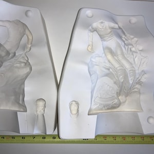May include: Two white plaster molds depicting figures in motion. One mold shows a skier, the other a figure on a rocky surface. Each mold has a small, separate piece. A yellow measuring tape is at the bottom.