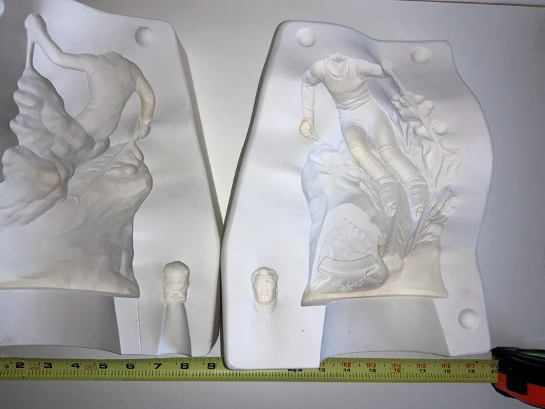 May include: Two white plaster molds depicting figures in motion. One mold shows a skier, the other a figure on a rocky surface. Each mold has a small, separate piece. A yellow measuring tape is at the bottom.