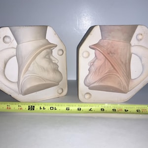 May include: Two ceramic molds, each featuring a profile of a man wearing a hat and a beard. The molds are white and have a handle shape. A yellow measuring tape is placed below the molds, showing measurements in inches.