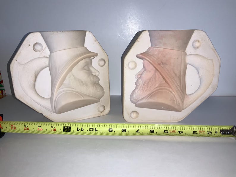 May include: Two ceramic molds, each featuring a profile of a man wearing a hat and a beard. The molds are white and have a handle shape. A yellow measuring tape is placed below the molds, showing measurements in inches.