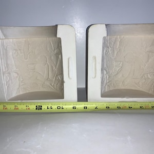 May include: Two off-white ceramic molds with detailed relief designs. Each mold has a rectangular shape with a curved interior and handles on the sides. A yellow measuring tape is placed below the molds, showing measurements in inches.