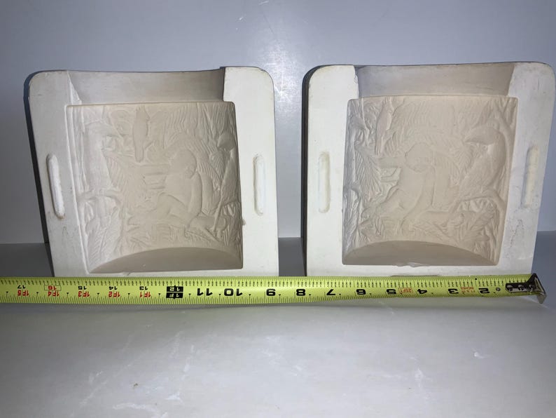 May include: Two off-white ceramic molds with detailed relief designs. Each mold has a rectangular shape with a curved interior and handles on the sides. A yellow measuring tape is placed below the molds, showing measurements in inches.