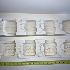 May include: Two white ceramic molds, each holding four mugs. The top mold features mugs with a beard and scarf design. The bottom mold has mugs with a Santa Claus face design. A yellow measuring tape is at the bottom.