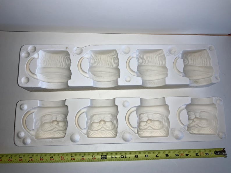May include: Two white ceramic molds, each holding four mugs. The top mold features mugs with a beard and scarf design. The bottom mold has mugs with a Santa Claus face design. A yellow measuring tape is at the bottom.