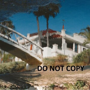 May include: A waterfront scene with a white building featuring a red roof and a bridge reflected in the water. The sky is blue, and palm trees and other greenery surround the building. The image has a slight reflection effect.