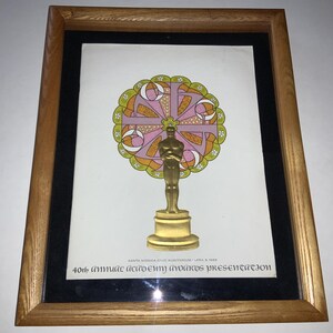 May include: Framed print of the 40th Annual Academy Awards presentation. The print features a gold Oscar statue in front of a colorful, circular design. The frame is made of light brown wood with a black matting.