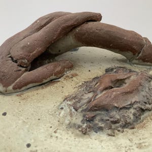 May include: A ceramic sculpture with a hand-shaped handle. The handle is a reddish-brown color, and the base is a light beige with dark brown speckles. The sculpture has a rough, textured surface.