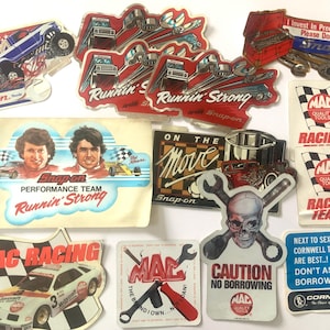 May include: A collection of assorted automotive and tool-themed stickers featuring Snap-on, Mac Tools, and Cornwell brands. Designs include race cars, a skull with crossed wrenches, and humorous text such as I Invest In Precious Metals Please Don't Ask to Borrow.