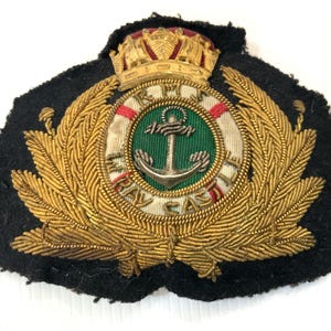 May include: A vintage naval cap badge featuring a gold crown, a central emblem with an anchor, and the words "Royal Naval" in a circular design. The badge is surrounded by gold-colored laurel leaves on a black felt backing.