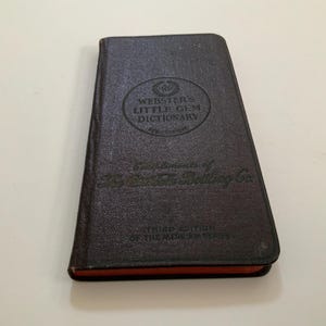 May include: A dark brown, rectangular pocket dictionary titled Webster's Little Gem Dictionary. The cover features embossed text including Third Edition of the Merriam Series and Complements of The Coca-Cola Bottling Co. The page edges are orange.