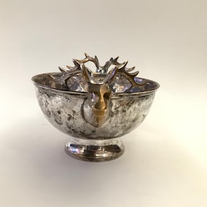Puede incluir: Cuenco de metal plateado con base de pedestal y diseño de cabeza de ciervo. El cuenco tiene un acabado deslustrado y la cabeza de ciervo es de color dorado. Las astas se extienden sobre el borde del cuenco.