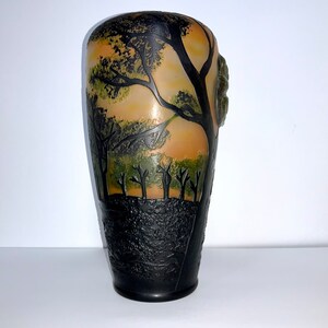 May include: A tall, black glass vase with a textured surface. The vase features a design of trees and a sunset in shades of green, brown, and orange.