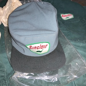 May include: A green work uniform with a blue and black Sinclair cap. The cap has a green dinosaur logo with the word Sinclair in red.