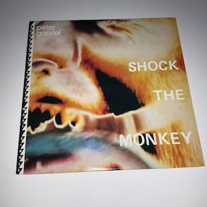 May include: A CD single for Peter Gabriel's "Shock the Monkey" featuring a close-up, abstract image of a face in shades of orange, yellow, and black. The title is printed in white block letters.