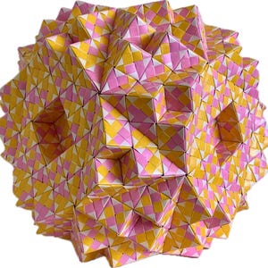 May include: A geometric, spherical sculpture composed of folded paper. The sculpture features a repeating pattern of pink, yellow, and white diamond shapes, creating a complex and textured surface. The overall form is a three-dimensional sphere.