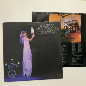 May include: Album cover for Stevie Nicks' Bella Donna. The album cover features a woman in a flowing lavender dress holding a white dove against a black background. The album title and artist name are in white script.