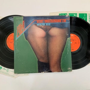 May include: Vinyl record album featuring the Velvet Underground Live with Lou Reed. The album cover has a green background with an illustration of a person's legs and buttocks. Two black vinyl records are visible.