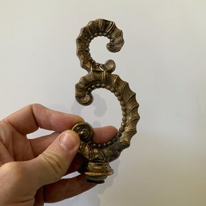 May include: Ornate bronze-colored decorative hardware with a scrolled design. The hardware has a textured surface and a detailed, swirling pattern. The piece is held in a hand against a plain white background. The hardware is likely for furniture or decor.