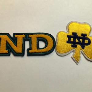 Può includere: Due toppe ricamate: una con le lettere "ND" in oro con bordo verde e un trifoglio dorato con "ND" in blu navy. Queste toppe sono probabilmente per abbigliamento o accessori.