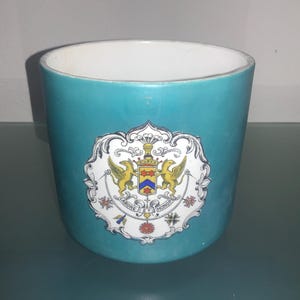 May include: A teal ceramic mug with a white crest featuring a coat of arms with two golden birds, a blue and yellow shield, and the words "Labore et Honore".