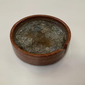 May include: A round, brown ceramic bowl with a rough texture. The bowl is filled with clear, crystalline material, possibly ice or decorative glass. The bowl has a simple, rustic design.
