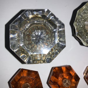 May include: Close-up of several vintage glass doorknobs. The top doorknob is clear and octagonal, with a starburst design in the center. Other doorknobs are visible, including a clear, textured one and several amber-colored ones. The doorknobs are arranged on a white surface.