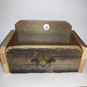 May include: A rustic wooden box with a distressed finish. It features a brass handle and a small, round knob with a floral design. The box is constructed from reclaimed wood, showcasing natural grain variations and a vintage aesthetic.