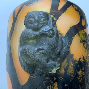 May include: A vintage glass vase with an orange and yellow background and a black silhouette of a tree. A black and green three-dimensional lemur is sculpted on the vase.