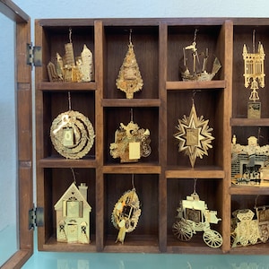 May include: A wooden display case with twelve gold-colored Christmas ornaments. The ornaments are arranged in a grid pattern and feature various designs, including a Christmas tree, a star, a house, and a ship.