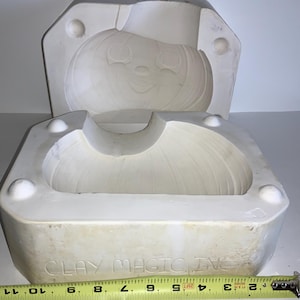 May include: A white ceramic mold for creating a pumpkin-shaped item. The mold is in two pieces, with a smiling pumpkin face design. The base of the mold has the text "CLAY MAGIC INC" and a measuring tape is placed below the mold.