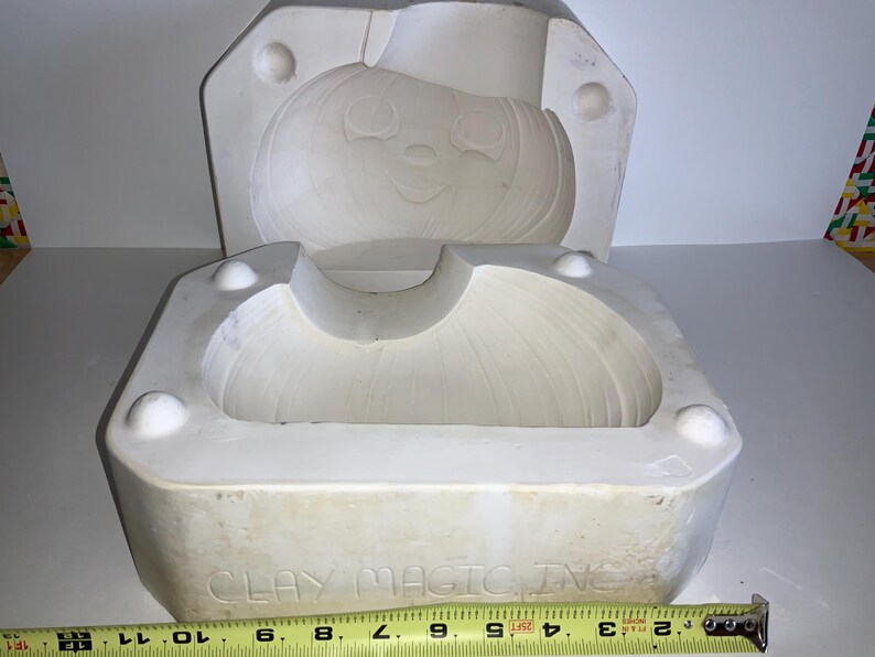 May include: A white ceramic mold for creating a pumpkin-shaped item. The mold is in two pieces, with a smiling pumpkin face design. The base of the mold has the text "CLAY MAGIC INC" and a measuring tape is placed below the mold.