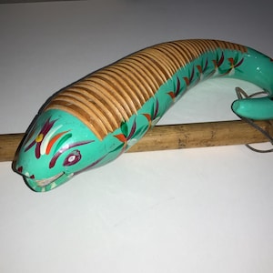 May include: A hand-painted wooden güiro shaped like a snake. The instrument features a turquoise head and tail, with a ribbed wooden body. Decorative floral accents in red, orange, and green adorn the body. A wooden stick rests beneath the instrument.