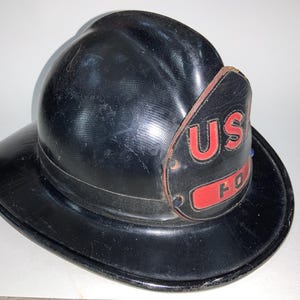 Könnte beinhalten: Schwarzer Feuerwehrhelm mit breiter Krempe. Der Helm hat eine strukturierte Oberfläche und ein Lederschild mit den roten Buchstaben "US". Das Wort "FO" ist ebenfalls auf dem Schild zu sehen.