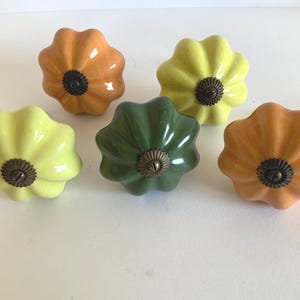 May include: Five ceramic cabinet knobs in shades of yellow, orange, and green. Each knob has a flower-like shape with a dark metal center. These decorative knobs are suitable for furniture.