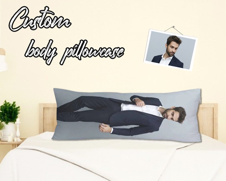 Op de afbeelding: Een grijze kussensloop voor een kussen voor het hele lichaam met een foto van een persoon in pak die erop ligt. De kussensloop ligt op een wit bed met een wit hoofdeinde. De tekst "Custom body pillowcase" staat op de muur achter het bed.