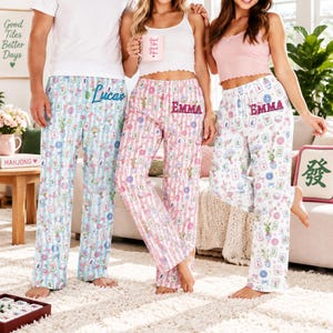 Könnte beinhalten: Drei Schlafhosen mit einem Muster aus Mahjong-Steinen und Blumen. Ein Modell mit blauen Streifen trägt den Namen Lucas, ein Modell mit rosa Streifen den Namen Emma und ein weißes Modell ebenfalls den Namen Emma. Jede Hose hat einen Gummibund.