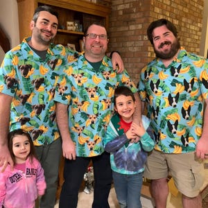 May include: Four people, three adults and one child, are wearing blue shirts with a banana and dog face pattern. The adults are smiling and the child is looking at the camera. The adults are wearing the shirts with the collars unbuttoned. The child is wearing a red shirt under a blue tie-dye hoodie. The adults are standing in front of a brick wall.