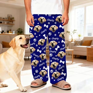May include: Blue pajama pants with a repeating pattern of a dog's face, paw prints, and bones. The word "Charly" is written in white script throughout the design. The pajama pants are worn with brown slippers.