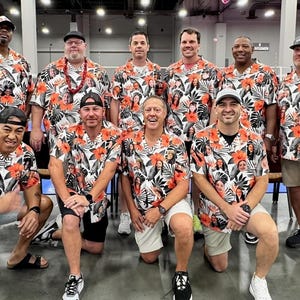 May include: A group of men wearing white and orange Hawaiian shirts with various faces printed on them. The shirts have a tropical print with palm leaves and flowers. The men are smiling and posing for a photo.