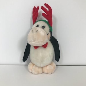 Könnte beinhalten: Ein Plüschpinguin mit roter Fliege, roten Geweih und grünem Hut. Der Pinguin hat einen weißen Körper, schwarze Flügel und beige Füße und Gesicht. Das Stofftier ist für die Weihnachtszeit gedacht.