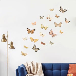 Könnte beinhalten: Eine weiße Wand mit einer Sammlung von Schmetterlings-Wandaufklebern in verschiedenen Brauntönen, Beige und Rosa. Die Schmetterlinge sind in einem zufälligen Muster angeordnet und schaffen ein verspieltes und elegantes Design.