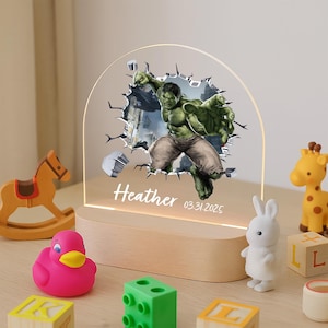 May include: A personalised night light with the Hulk breaking through a wall design. The light has a wooden base and an arched acrylic panel with the name "Heather" and the date "03.31.2025". Children's toys surround the light.