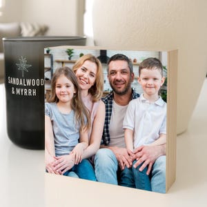 May include: A rectangular wooden photo block displaying a colour photograph of a family of four, including two adults and two children, sitting together. Next to the block is a black candle holder with the text SANDALWOOD & MYRRH printed on it.