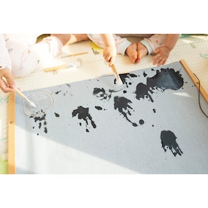 May include: A grey canvas stretched on a wooden frame with black paint splatters. Two wooden handled paintbrushes are on the canvas. A small clear container with a lid holds paint.