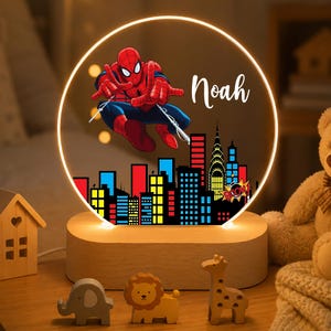 May include: A circular LED night light featuring a graphic of Spider-Man in a red and blue suit leaping above a colourful city skyline. The name Noah is printed in white script. The light is mounted on a light-coloured wooden base.