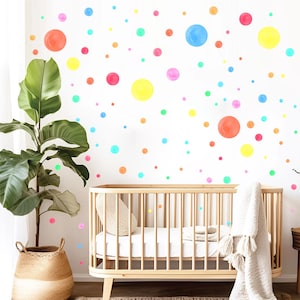 May include: A nursery interior featuring a light wood cot, a large potted plant, and a wall adorned with colourful polka dots. The dots are in shades of orange, pink, yellow, blue, and green, creating a playful and cheerful atmosphere.